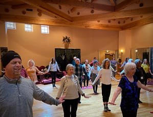 Group of participants engaged in a Chen Chi movement practice in a warm and inviting Kiva setting, with natural light illuminating the wooden beams and serene atmosphere.