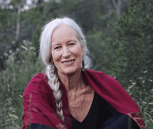 Portrait of Cynthia Jurs sitting outdoors. She is wearing a dark top and a rich red shawl draped over her shoulders, complementing her gentle smile and twinkling eyes, which convey a sense of wisdom and calm. The lush greenery in the background enhances the peaceful and natural atmosphere, and reflecting her connection to the environment.