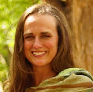 A portrait of Sarah, a woman with long brown hair and a warm smile. She is outdoors, wrapped in a green shawl, with a backdrop of soft greenery and tree bark, creating a natural and serene atmosphere.