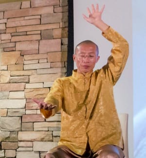 Master Mingtong Gu seated and demonstrating a Wisdom Healing Qigong practice, guiding energy flow with focused intention against a backdrop of a stone-accented wall.