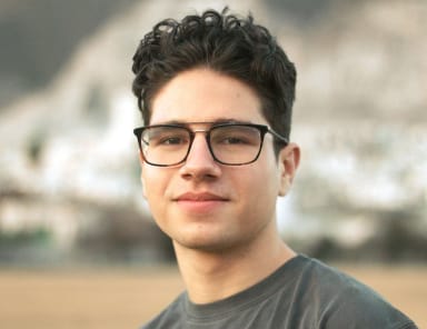 Portrait of a young man with curly hair and glasses, wearing a casual gray shirt, standing outdoors with a blurred natural background.