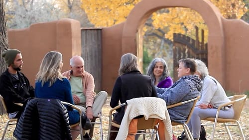 Group of people sitting in a circle outdoors under autumn trees near an adobe archway, engaging in a reflective or healing discussion, promoting community and connection in a peaceful natural setting.