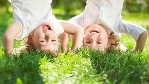 Two joyful children playing upside-down on lush green grass, laughing and enjoying a sunny day outdoors.