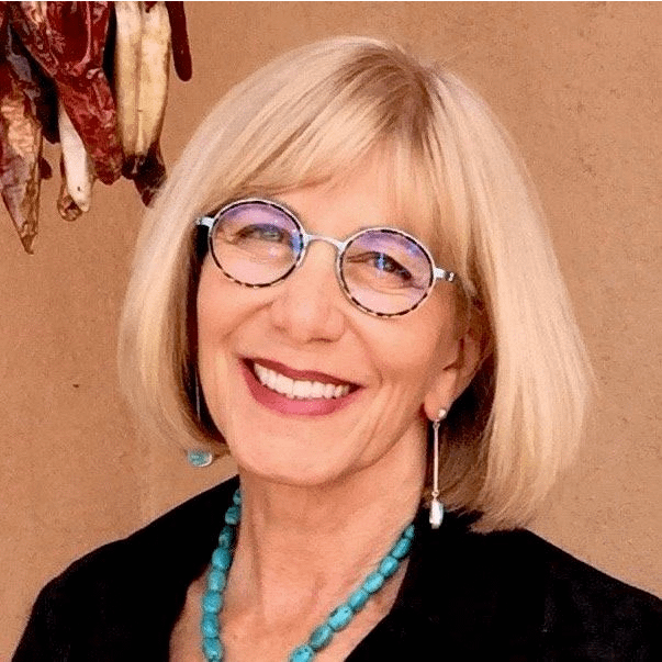 A cheerful woman with short blonde hair, wearing round glasses, turquoise jewelry, and a black top, standing against an adobe wall with dried red chilies in the background, radiating warmth and charm.