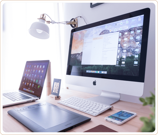 Modern workspace setup featuring an iMac desktop, a laptop, a smartphone, and a graphic tablet on a well-lit desk with an inspirational 'Never Settle' card.