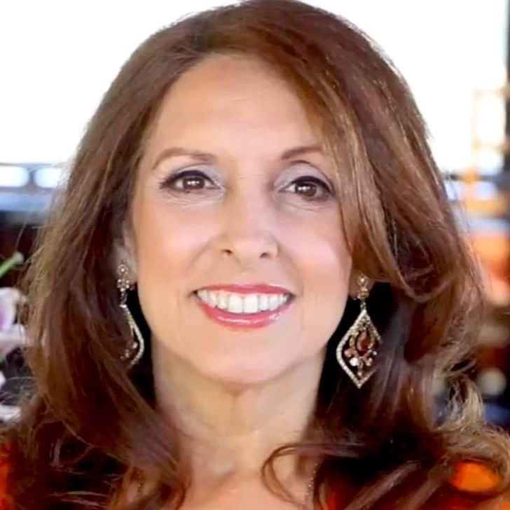 A smiling woman with long, wavy brown hair, wearing elegant drop earrings and a vibrant top, exuding confidence and warmth in a softly lit indoor setting.