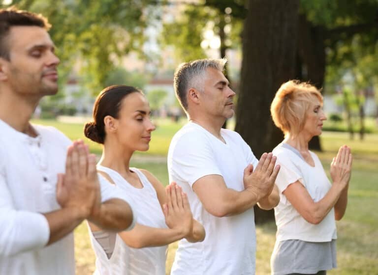 Group of diverse individuals practicing mindfulness meditation outdoors in a serene park setting, with hands in a prayer position and wearing white clothing, symbolizing peace and connection to nature