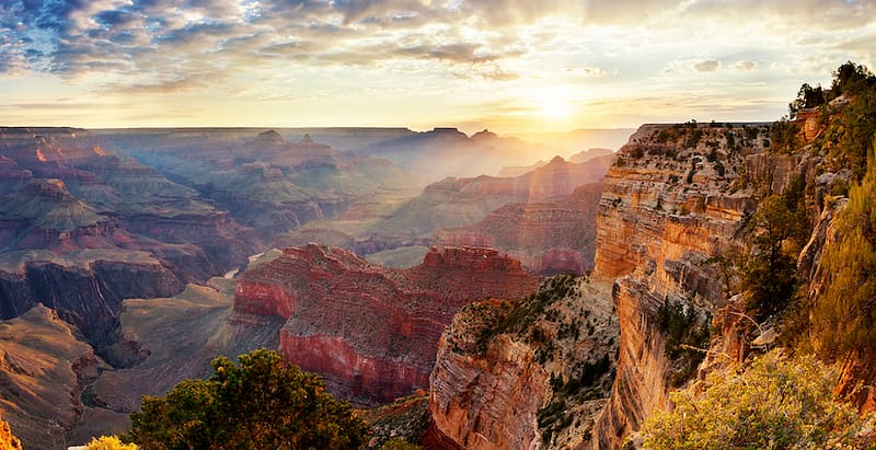 A panoramic view of the Grand Canyon at sunrise, with the sun casting a golden glow across the layered red and orange canyon walls. The vast expanse of the canyon is highlighted by dramatic shadows and light, revealing its immense depth and rugged beauty. The sky is partially cloudy, creating a soft, warm light that enhances the breathtaking landscape, while a few green trees on the canyon rim add a touch of color to the rocky terrain.