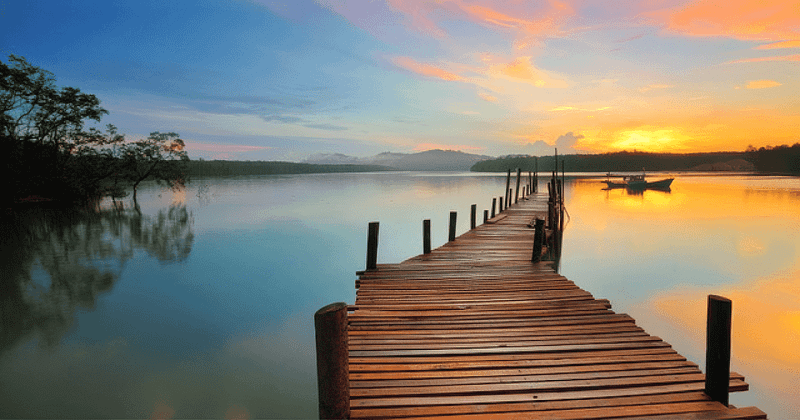 Serene early morning scene at a calm lake, showing a wooden dock stretching out towards the water with a small boat moored nearby. The sky is a canvas of soft oranges and pinks, reflecting on the still lake surface, embodying the tranquility and meditative essence of Wisdom Healing Qigong and the concept of Pure Consciousness.