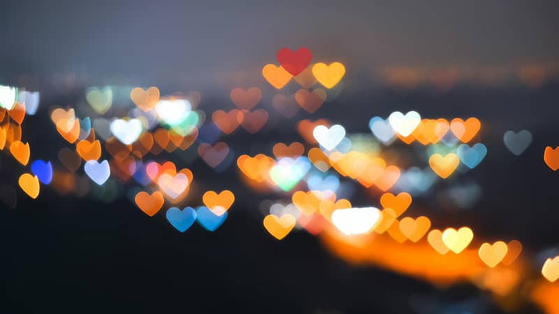 A vibrant night scene with colorful heart-shaped bokeh lights in various hues of orange, red, blue, and white, creating a dreamy and romantic atmosphere