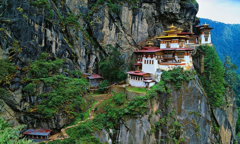 A breathtaking view of the Tiger's Nest Monastery (Paro Taktsang) clinging to the side of a steep cliff in Bhutan. This sacred Buddhist site is nestled among lush greenery and rugged rock faces, with steps winding up the mountain to its vibrant, ornate buildings. The red and gold roofs contrast strikingly against the natural rock, adding to the mystical atmosphere of this serene, remote setting amidst the Himalayan landscape.