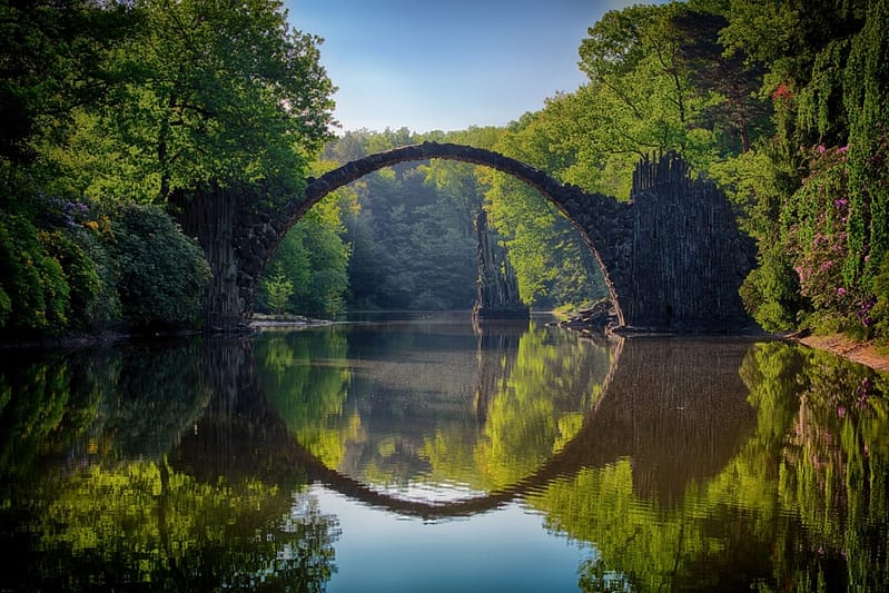 The image captures a picturesque stone arch bridge, known for its near-perfect circular reflection on the still water below, creating an illusion of a complete circle. The bridge is surrounded by lush green trees and dense foliage, with hints of flowering plants adding subtle colors to the scene. The soft lighting gives a mystical quality to the landscape, enhancing the natural symmetry and tranquility of the setting. The calm water, mirroring the bridge and the surrounding greenery, adds to the ethereal beauty, inviting a sense of peace and wonder.