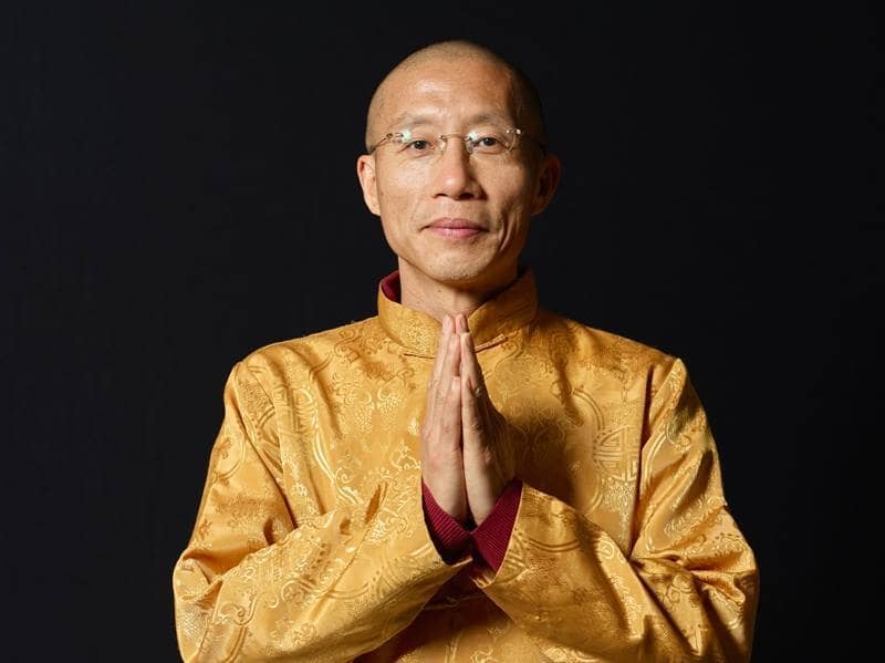 Master Mingtong Gu in traditional golden attire, offering a peaceful gesture with hands pressed together, representing wisdom and healing energy.