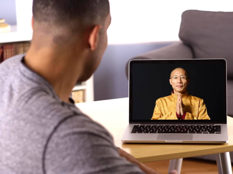 A man in a casual gray shirt engages in an online session on his laptop, featuring Master Mingtong Gu wearing a golden traditional outfit, teaching or guiding through a video on the screen in a home setting.