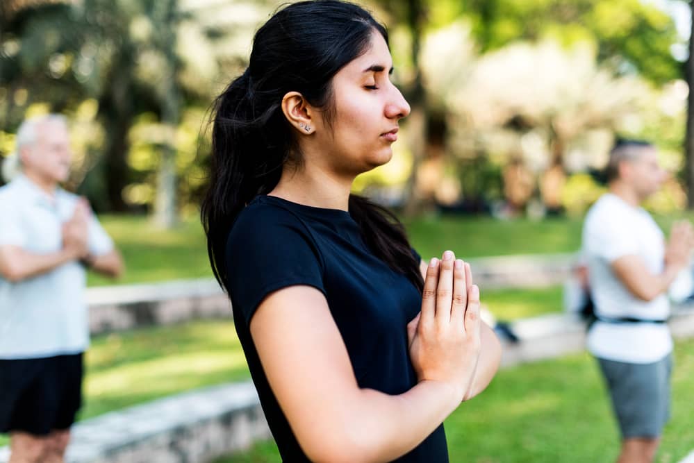 Peaceful young woman practicing mindfulness with hands in prayer position during an outdoor group meditation session, surrounded by lush greenery and soft natural light.